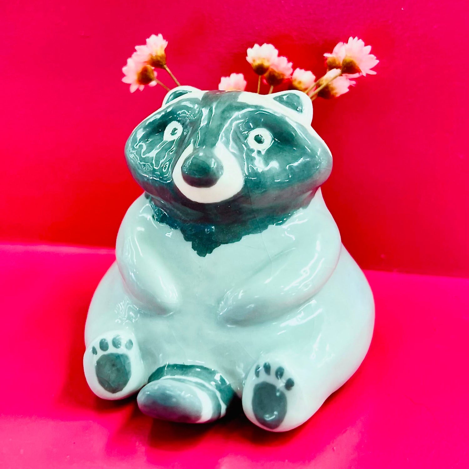 A miniature raccoon shaped vase coloured black and grey. Tiny pink flowers are placed in the vase. The vase is sitting on a bright pink shelf.