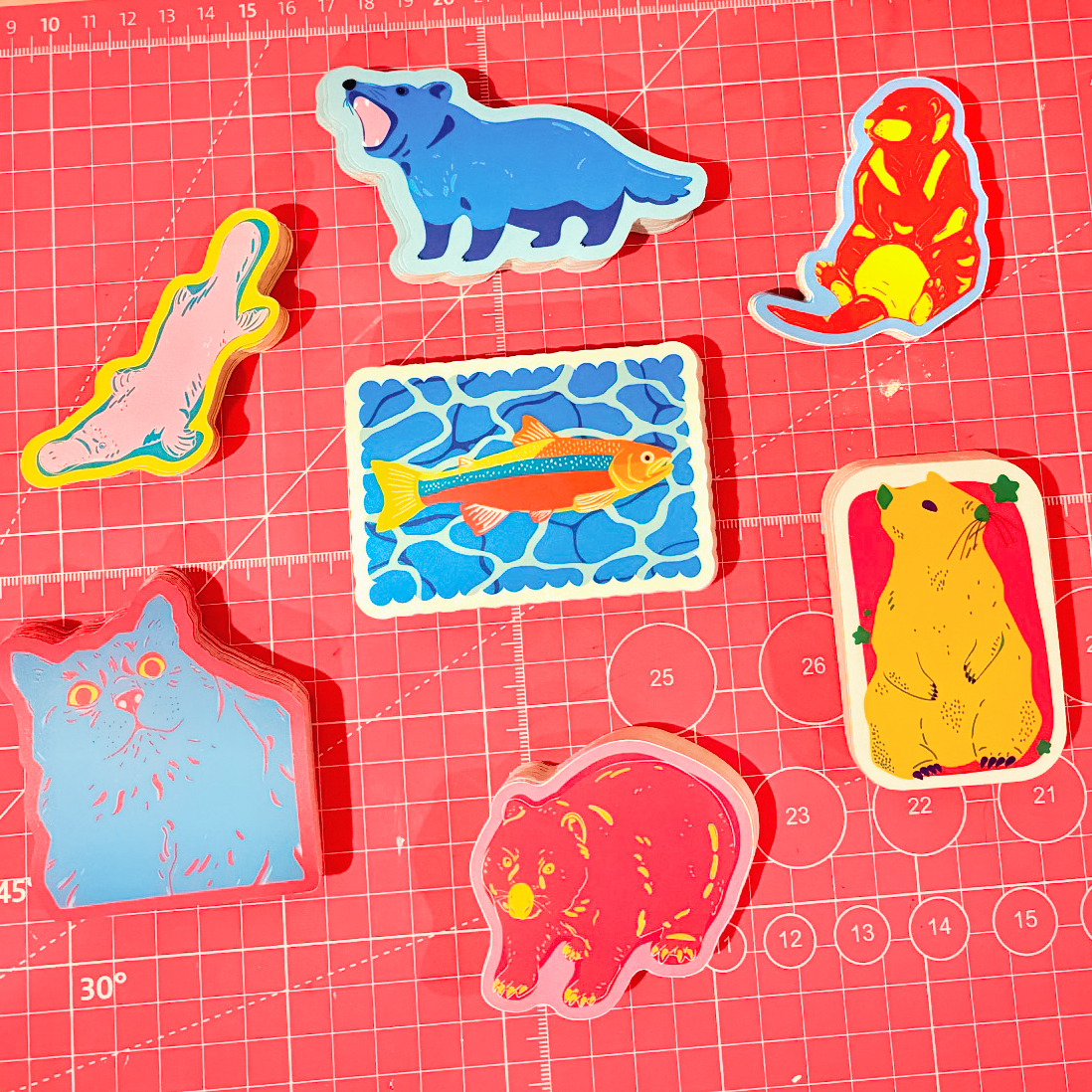 A collection of stickers including a cat, wombat, platypus, tasmanian devil, otter and marmot. Background is a pink checkered cutting mat.