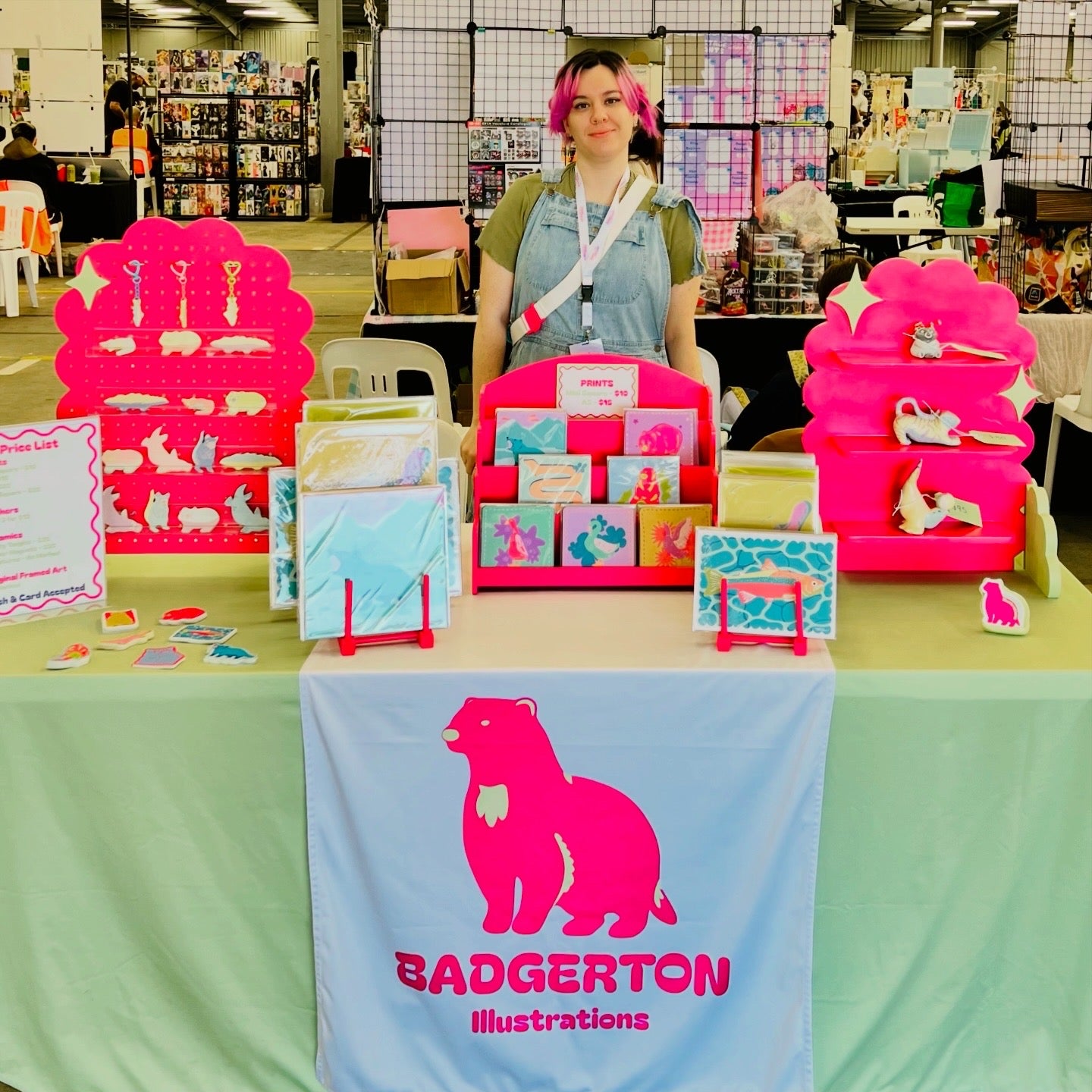 Jas standing behind her market table setup at an event. Featuring pale green table cloth, bright pink displays, and an assortment of prints, ceramics and stickers.