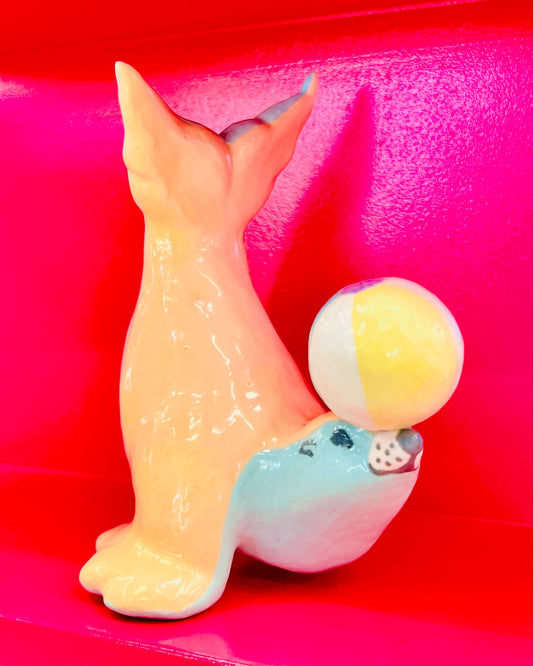 Ceramic sculpture of a blue and orange sea lion balancing a beach ball on its nose, sitting on a bright pink shelf.