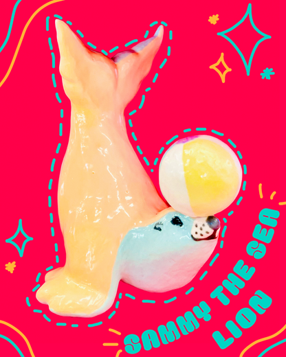 Ceramic sculpture of a blue and orange sea lion balancing a beach ball on its nose, with a bright pink background and decorative elements.